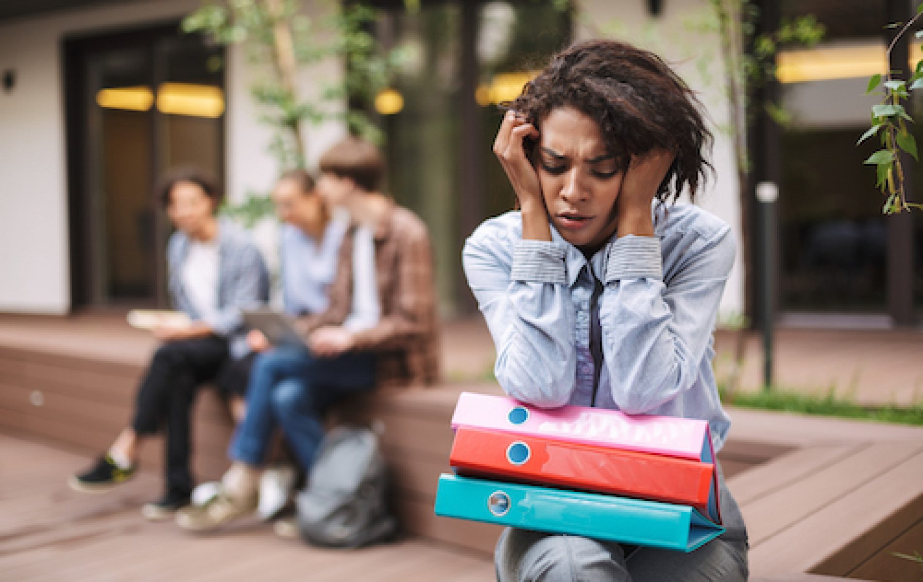 Portrait of upset lady sitting on bench with colorful folders on knees and sadly closing her eyes while spending time in courtyard of university