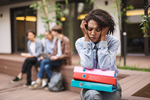 Portrait of upset lady sitting on bench with colorful folders on knees and sadly closing her eyes while spending time in courtyard of university